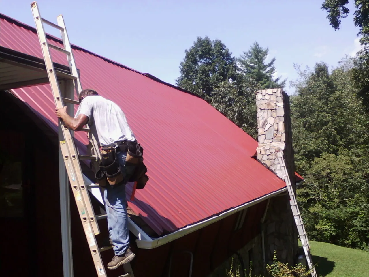 Roofer on a ladder beside a red metal roof and stone chimney — Soffit & Fascia Repair by Roofing Brothers in Universal City